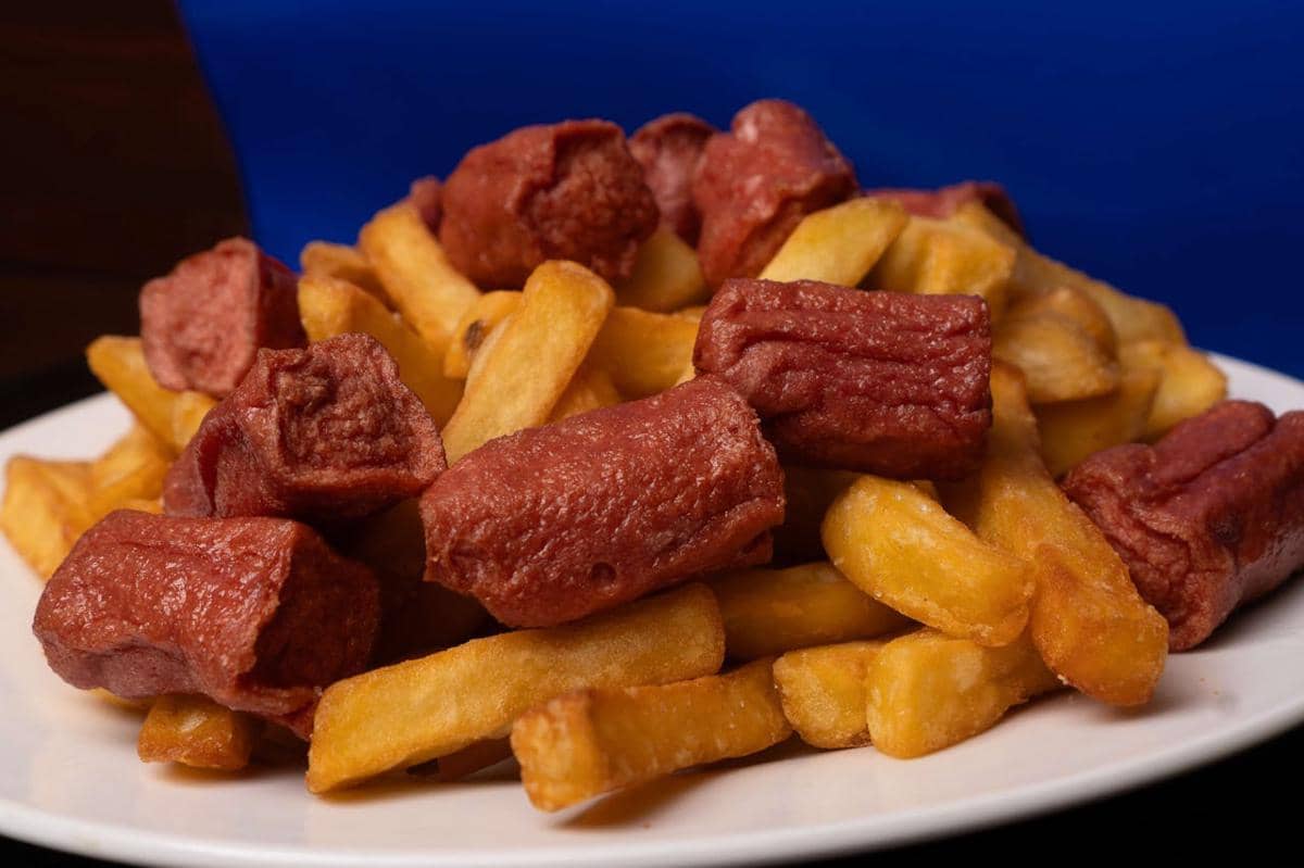 Tumpukan sosis ultraproses berwarna merah kecokelatan dan kentang goreng kuning keemasan di atas piring putih dengan latar biru.