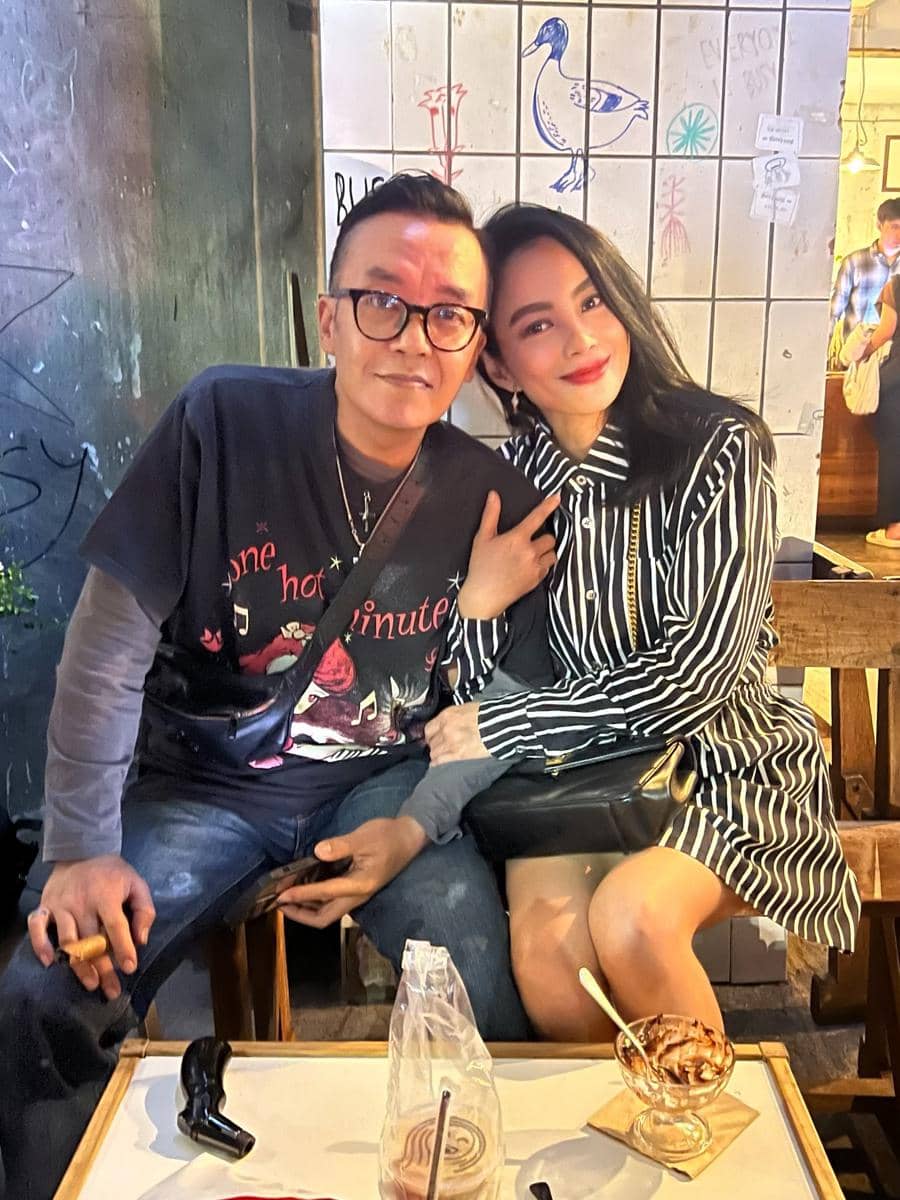 Seorang pria berkacamata duduk di samping seorang wanita berambut hitam dengan pakaian bergaris, keduanya tersenyum di sebuah kafe.