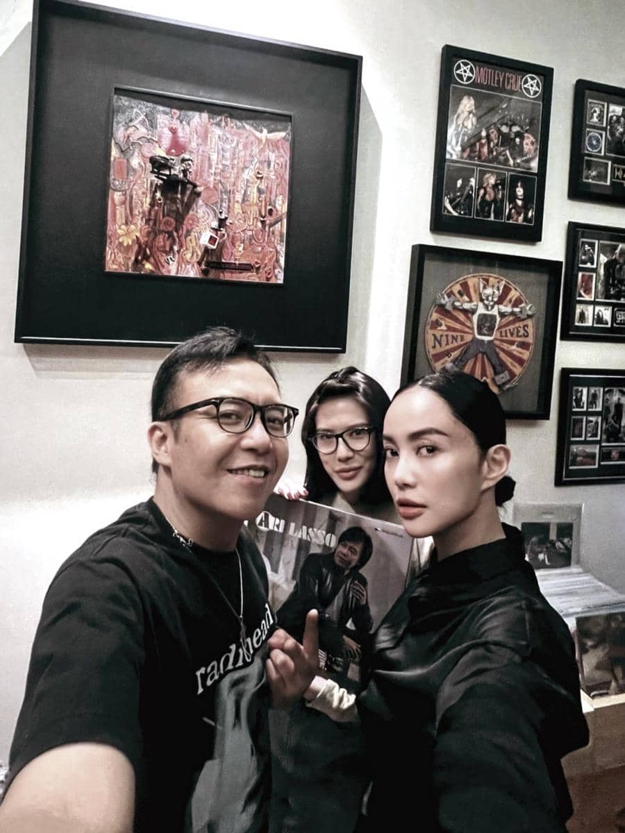 Tiga orang berpose bersama di ruangan dengan dinding penuh bingkai foto dan karya seni, salah satunya memegang piringan hitam bertuliskan Ari Lasso.