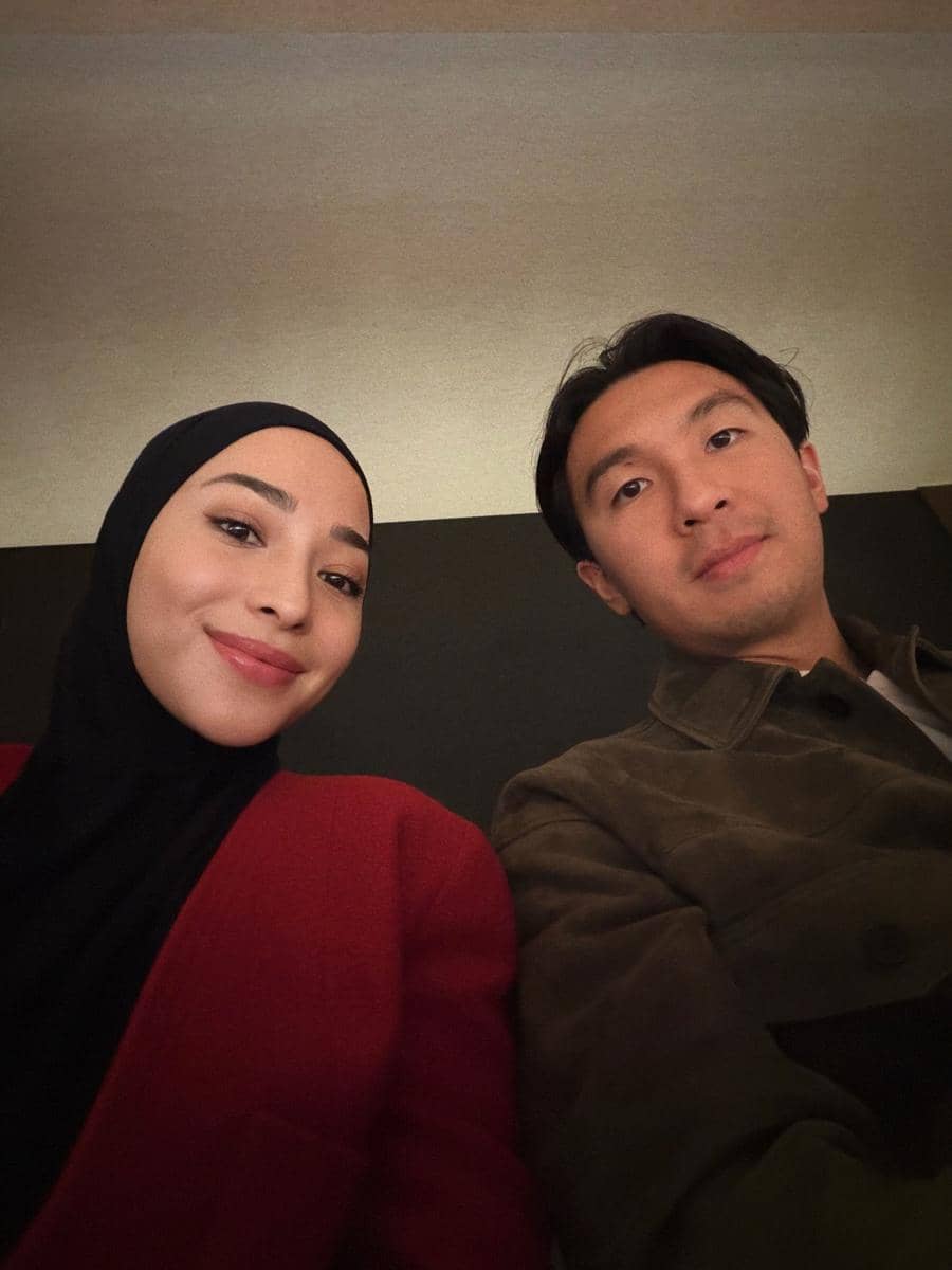 Seorang perempuan berhijab merah dan seorang pria berjaket cokelat tersenyum bersama dalam suasana hangat di dalam ruangan.