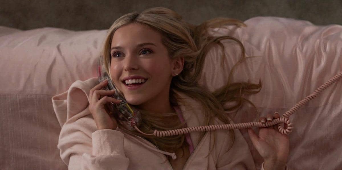 Seorang perempuan berambut pirang tersenyum sambil berbicara di telepon kabel berwarna merah muda, berbaring di tempat tidur dengan selimut pink.