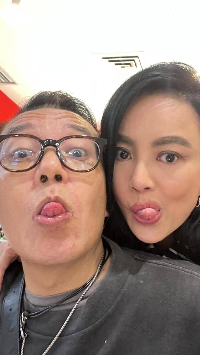Dua orang berpose selfie sambil menjulurkan lidah dengan ekspresi lucu di dalam ruangan bercat merah dan putih.