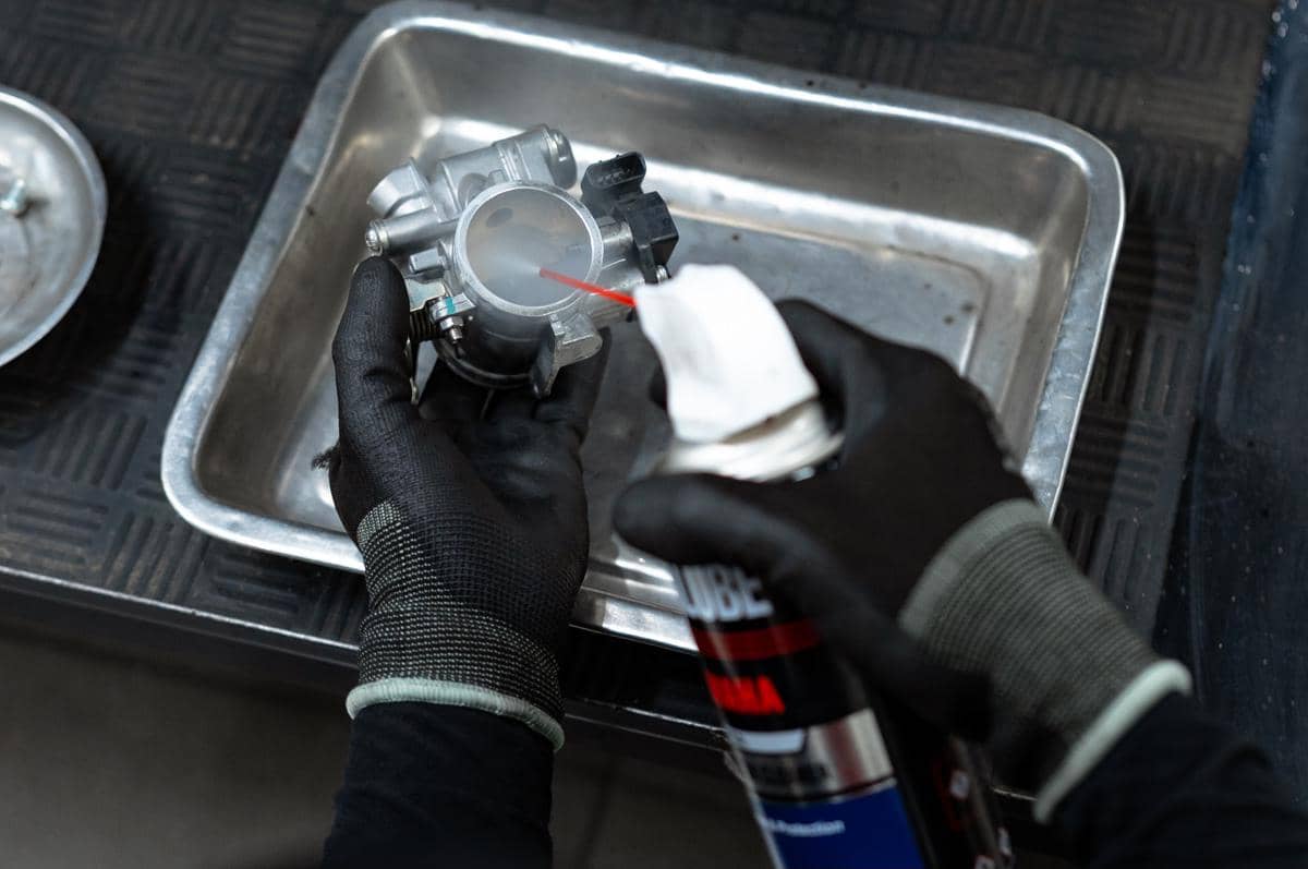 Teknisi menggunakan sarung tangan hitam sedang membersihkan throttle body motor dengan cairan pembersih di atas wadah logam.