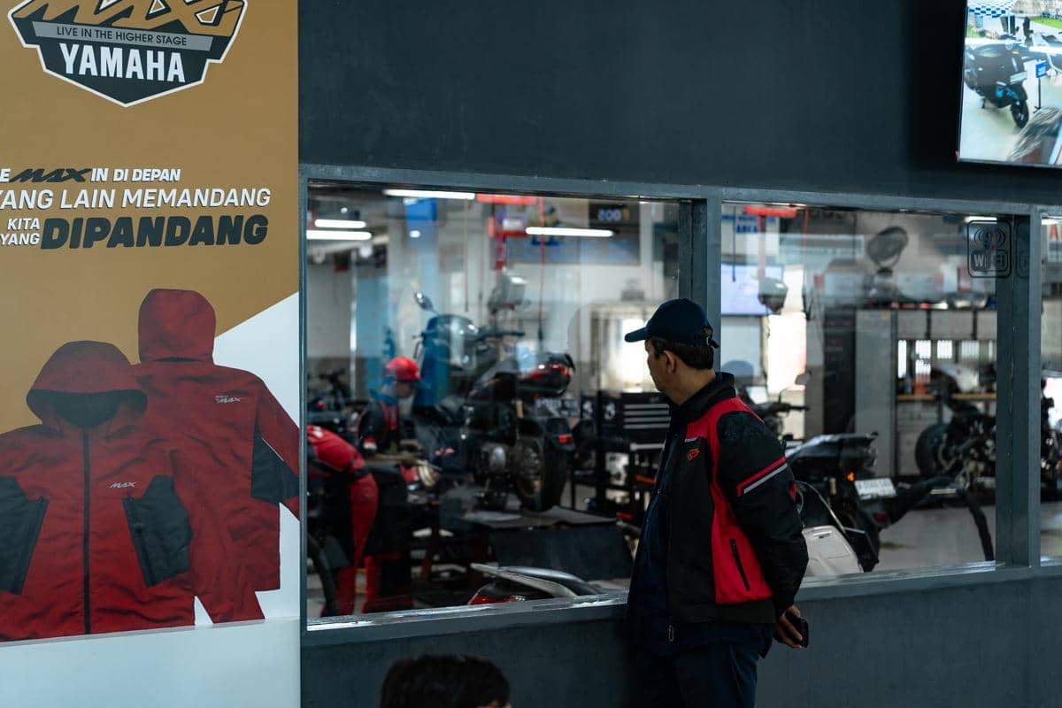 Seorang pria mengenakan jaket merah hitam melihat ke dalam bengkel Yamaha tempat mekanik bekerja memperbaiki motor.