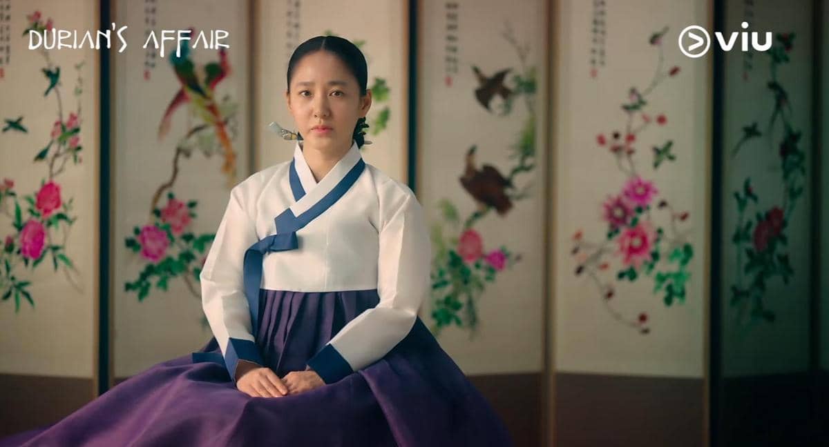Seorang wanita mengenakan hanbok tradisional Korea duduk dengan latar belakang sekat bergambar bunga dan burung dalam cuplikan drama Durian's Affair.