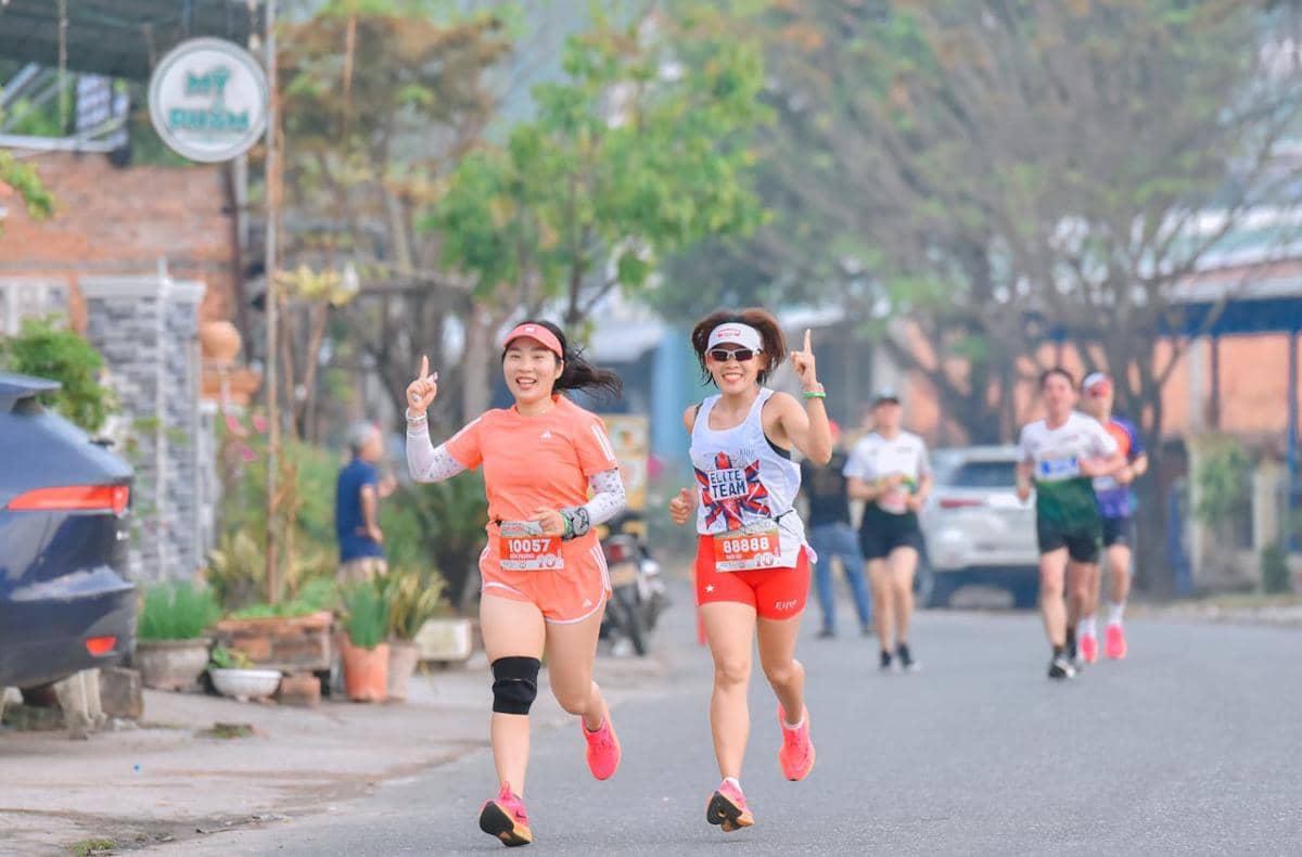 Dua pelari perempuan tersenyum sambil berlari di jalan perkotaan, mengenakan pakaian olahraga berwarna cerah dan nomor peserta lomba.