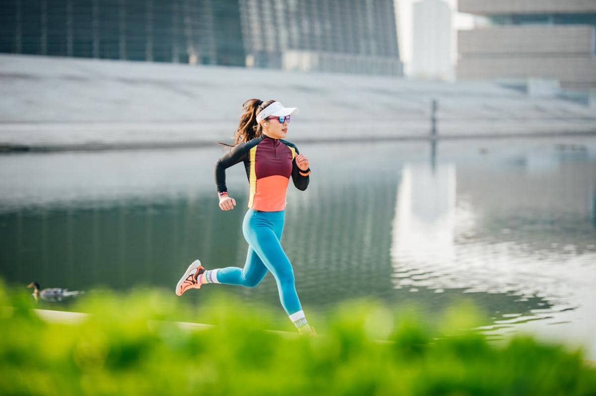 Seorang perempuan berlari cepat di tepi danau dengan latar gedung modern, mengenakan pakaian olahraga berwarna cerah dan kacamata hitam.