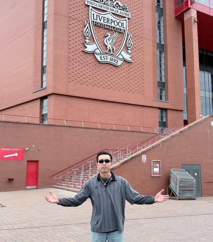 Seorang pria berdiri dengan tangan terbuka di depan dinding Stadion Anfield yang menampilkan logo besar Liverpool Football Club.
