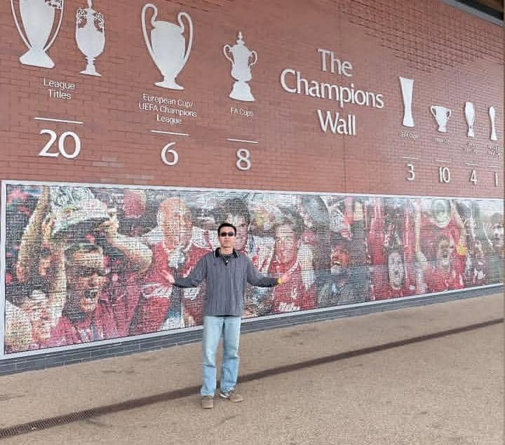 Seorang pria berpose di depan dinding The Champions Wall di Stadion Anfield yang menampilkan trofi dan pencapaian klub Liverpool.