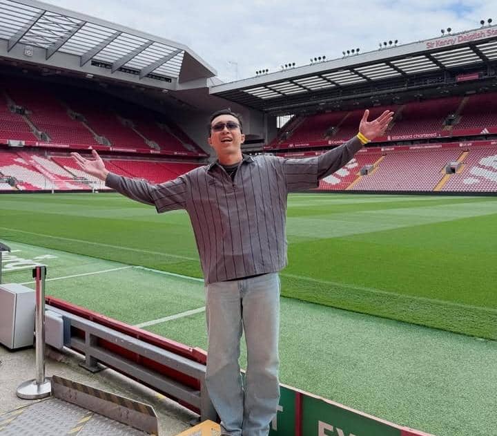 Seorang pria berdiri dengan tangan terbuka di tepi lapangan Stadion Anfield yang kosong dengan tribun berwarna merah di belakangnya.