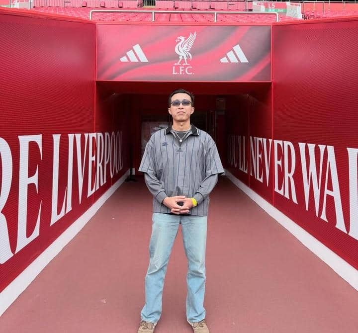 Seorang pria berdiri di lorong stadion Anfield dengan latar belakang logo Liverpool FC dan tulisan berwarna putih di dinding merah.