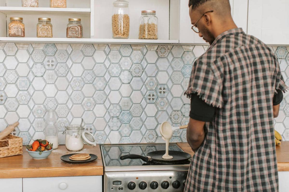 Seorang pria menuangkan adonan ke atas teflon di dapur dengan latar ubin bermotif heksagonal dan meja kayu berisi bahan masakan.