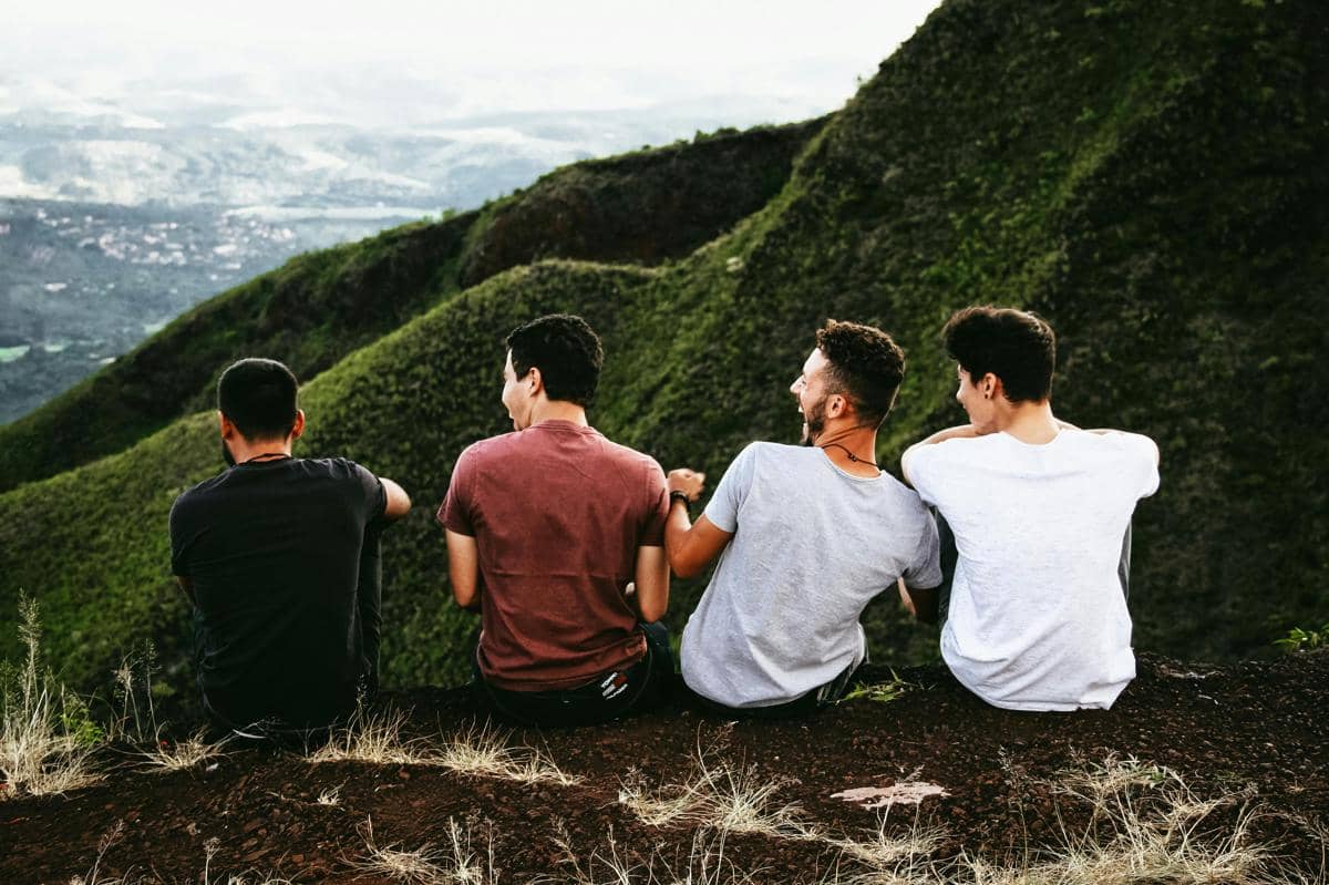 Empat sahabat duduk di tepi bukit hijau sambil tertawa bersama menikmati pemandangan alam yang indah di kejauhan.