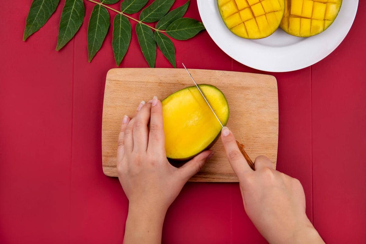 Ilustrasi tangan memotong mangga matang di atas talenan kayu dengan pisau dapur untuk menyiapkan potongan buah segar.