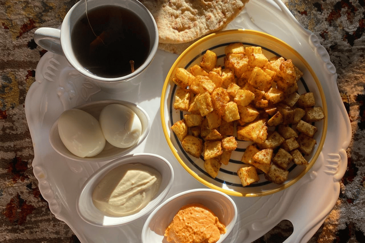 Sepiring kentang goreng berbumbu disajikan bersama telur rebus, roti pipih, saus, dan secangkir teh di atas nampan putih.