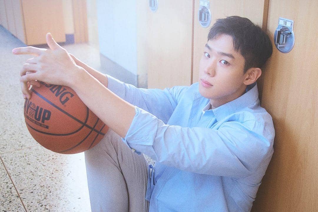 Seorang pria mengenakan kemeja biru muda duduk di lantai dekat loker sekolah sambil memegang bola basket dengan tatapan tenang.