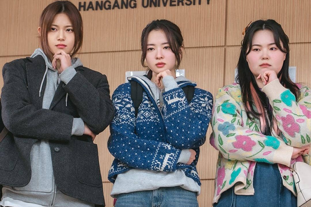 Tiga perempuan muda berdiri berdampingan dengan ekspresi serius di depan dinding bertuliskan Hangang University.