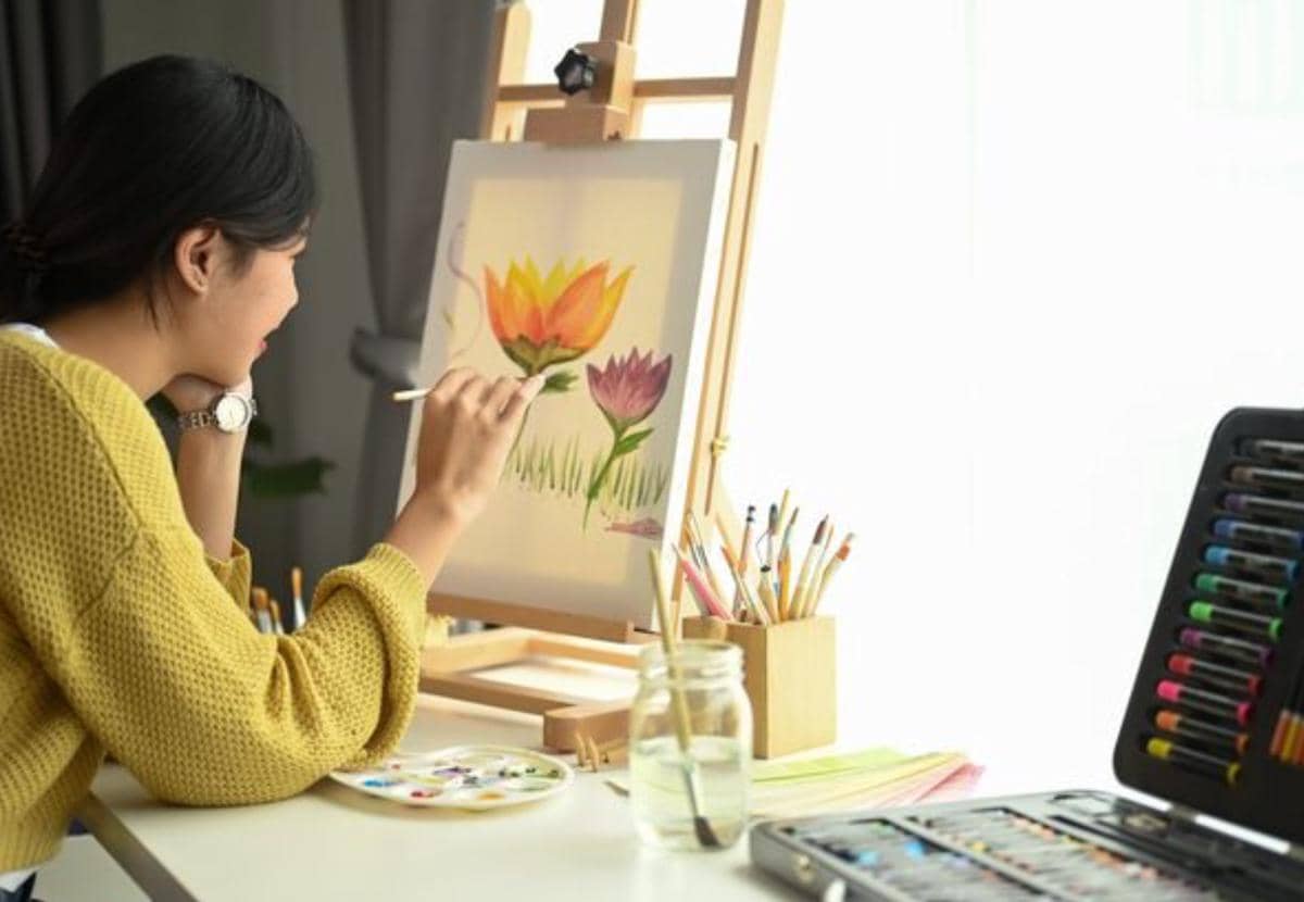Seorang ilustrator freelance mengenakan sweater kuning sedang melukis bunga di kanvas dengan cat air di meja kerja terang.