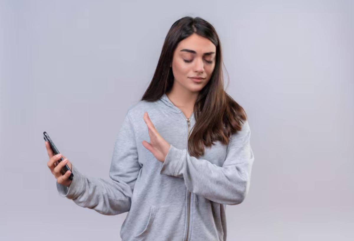 Seorang perempuan mengenakan hoodie abu-abu memegang ponsel sambil menolak dengan tangan, menggambarkan konsep digital detox.