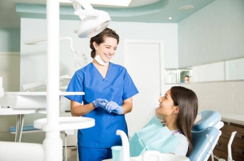 Seorang tenaga medis berpakaian biru tersenyum sambil berbicara dengan pasien perempuan yang duduk di kursi perawatan gigi di klinik.