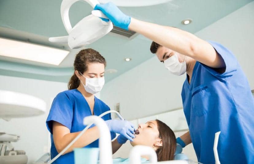 Dua tenaga medis mengenakan seragam biru dan masker sedang membersihkan karang gigi pasien wanita di klinik gigi modern.