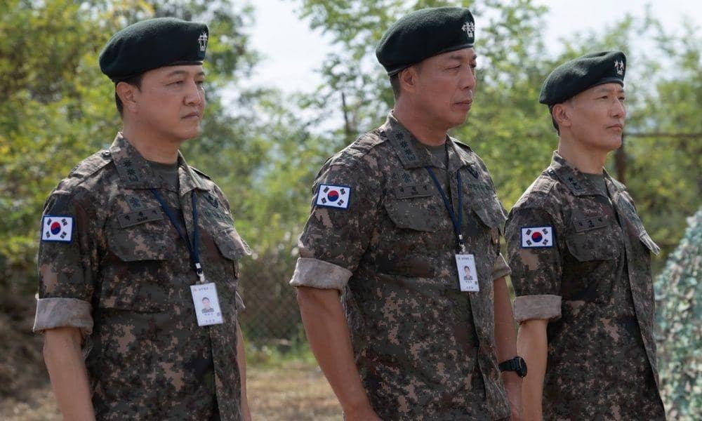 Tiga tentara mengenakan seragam militer Korea Selatan berdiri berbaris di luar ruangan dengan latar pepohonan dan langit cerah.