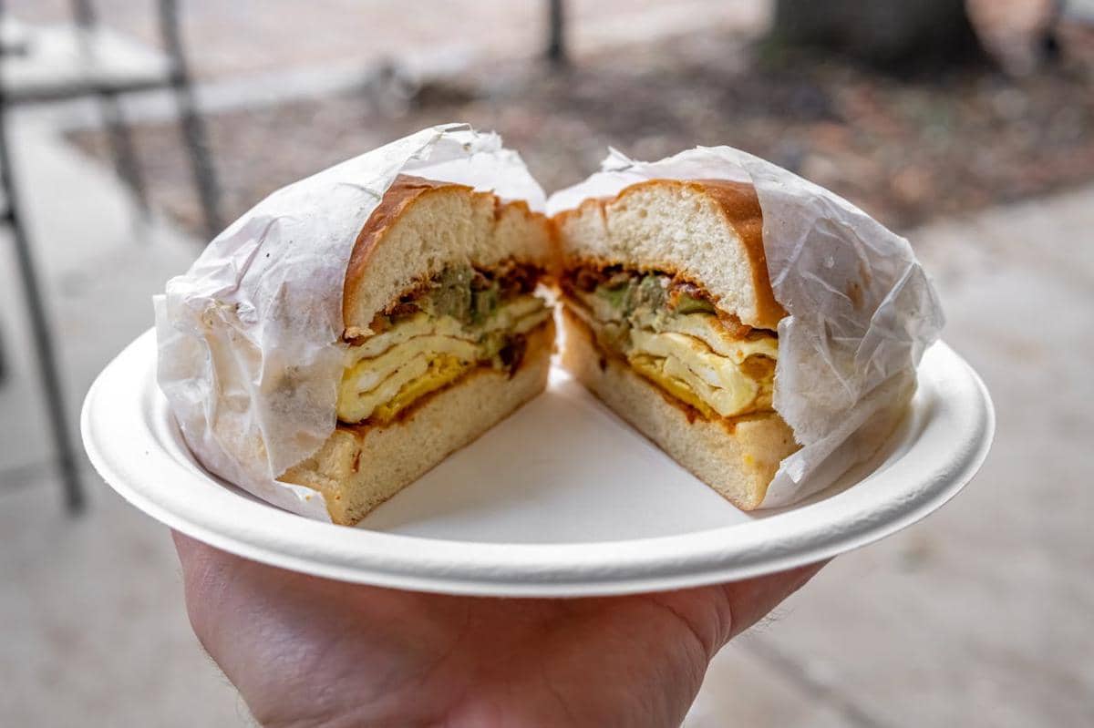 Roti sandwich berisi kentang dan telur disajikan di atas piring kertas putih, dibungkus sebagian dengan kertas pembungkus.