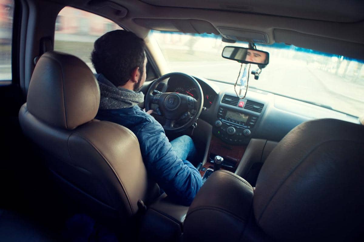 Seorang pria mengenakan jaket biru sedang mengemudi mobil di jalan dengan interior berwarna cokelat dan dashboard modern.