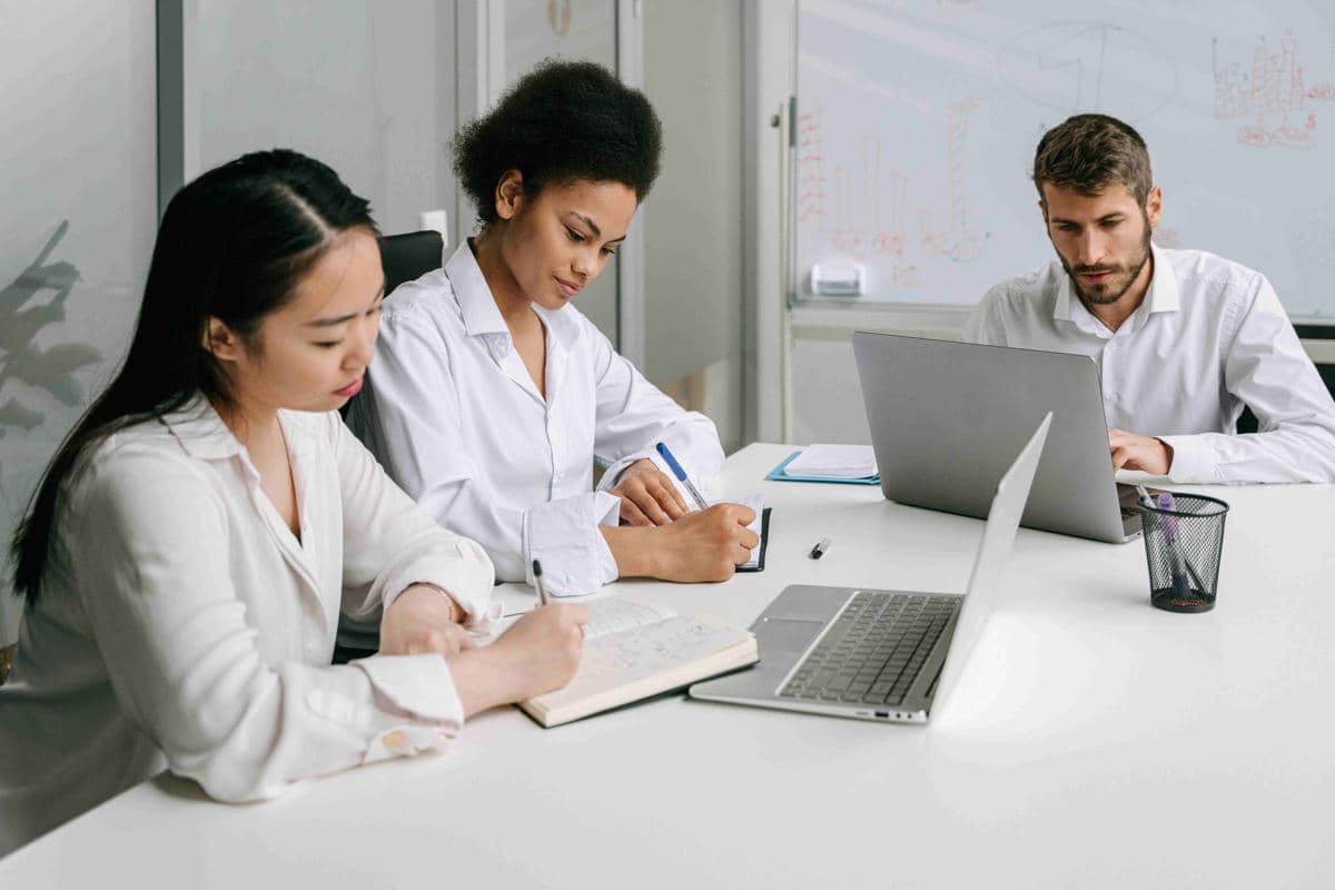 Tiga orang profesional duduk di meja kantor, dua menulis catatan di buku dan satu bekerja dengan laptop di depan papan tulis.
