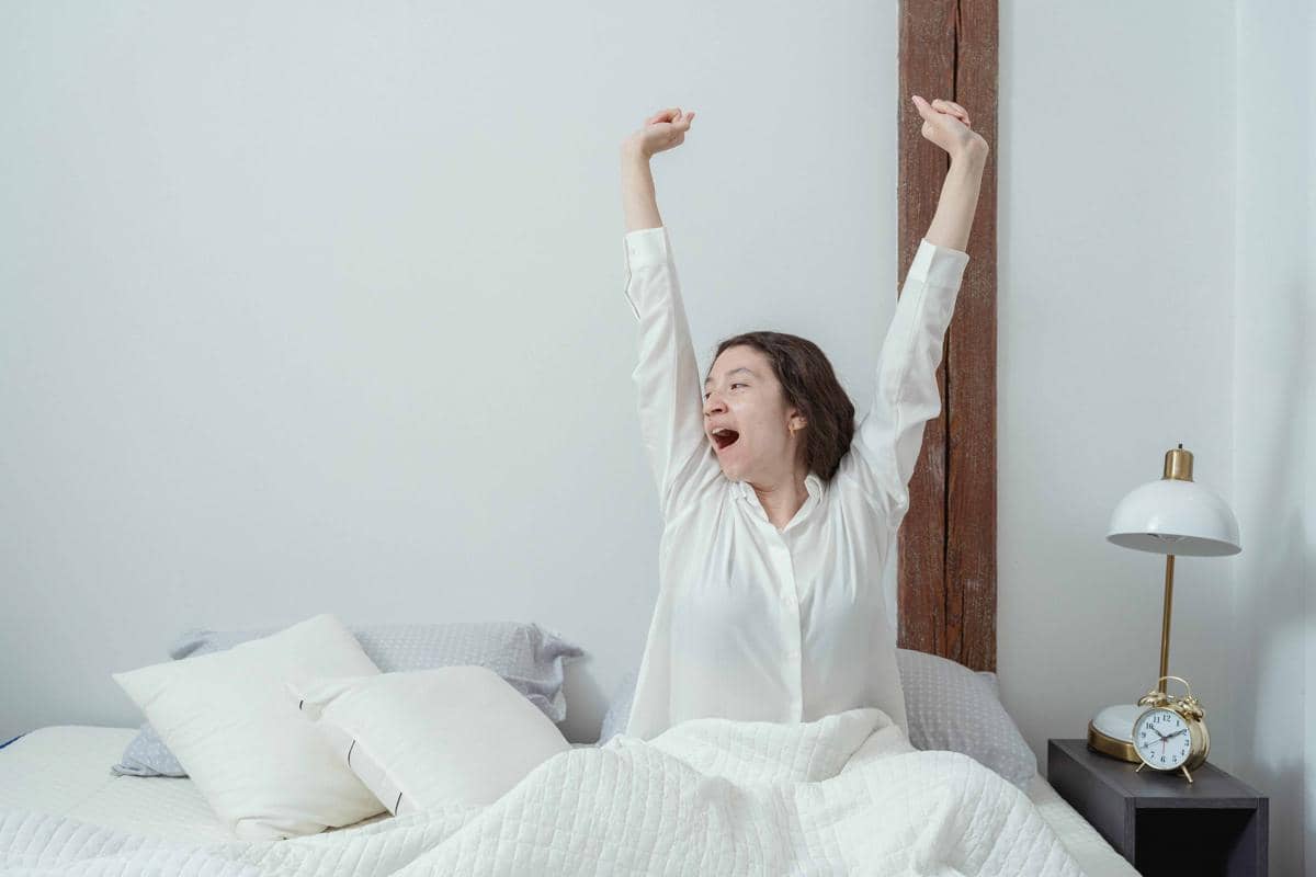 Seorang wanita mengenakan piyama putih sedang meregangkan tangan dan menguap di tempat tidur pada pagi hari di kamar minimalis.