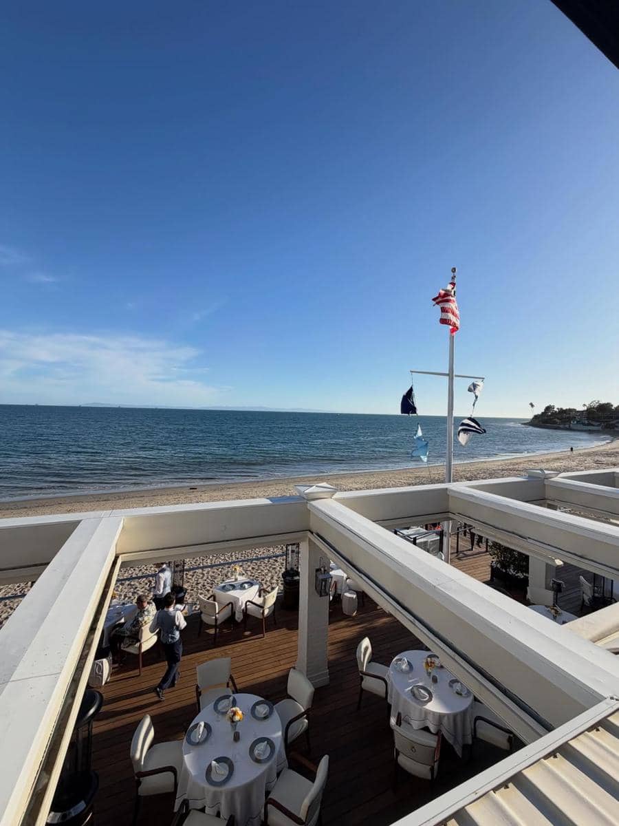 Pemandangan restoran tepi pantai di California dengan meja makan berlapis taplak putih dan bendera Amerika berkibar di bawah langit biru cerah.