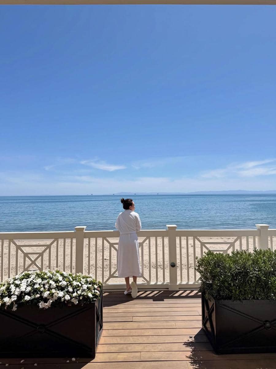 Seorang wanita mengenakan jubah putih berdiri di balkon kayu menghadap pantai berpasir dan laut biru di bawah langit cerah di California.