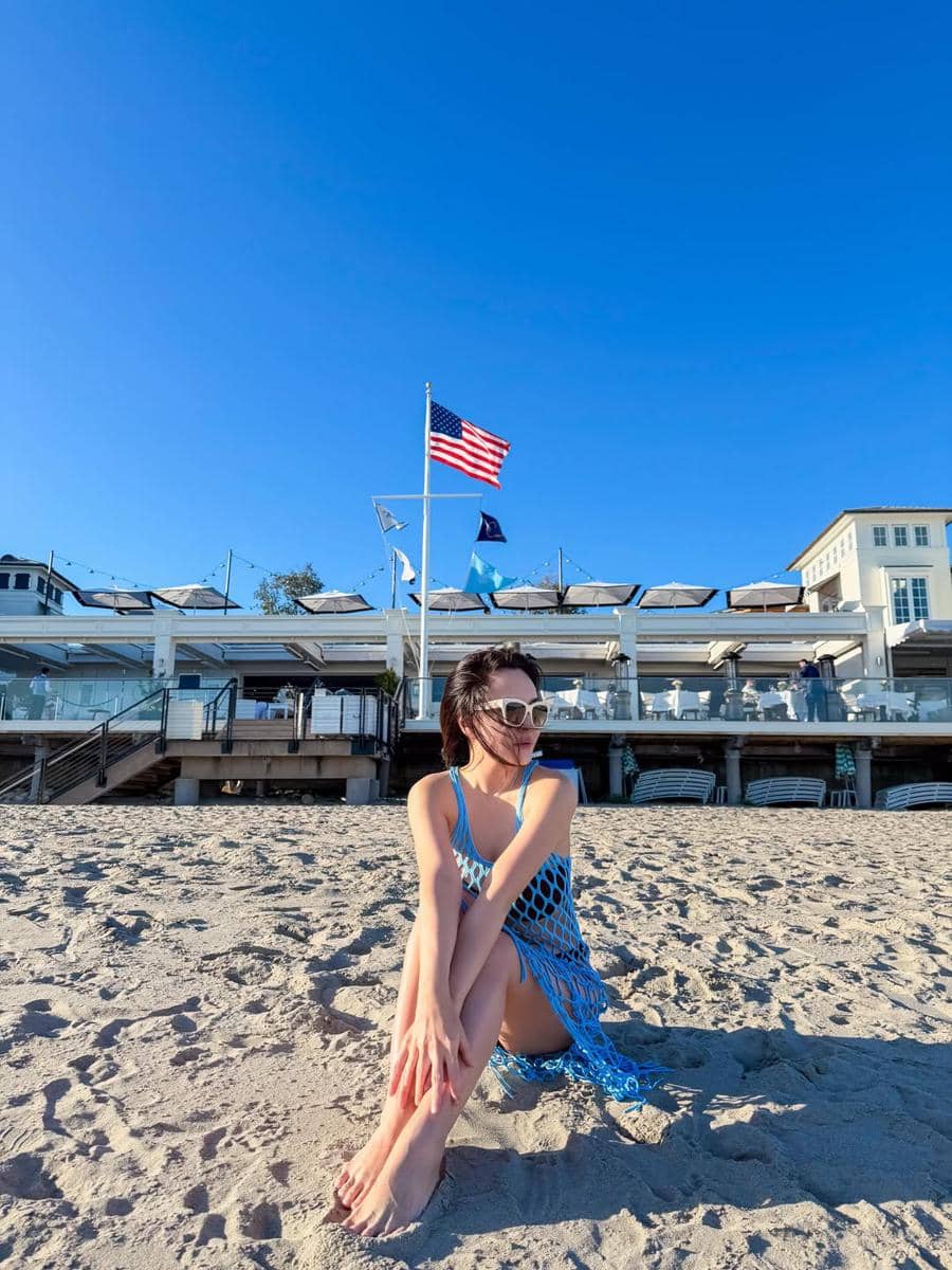 Seorang wanita duduk di pantai berpasir dengan latar belakang bangunan dan bendera Amerika Serikat di bawah langit biru cerah.