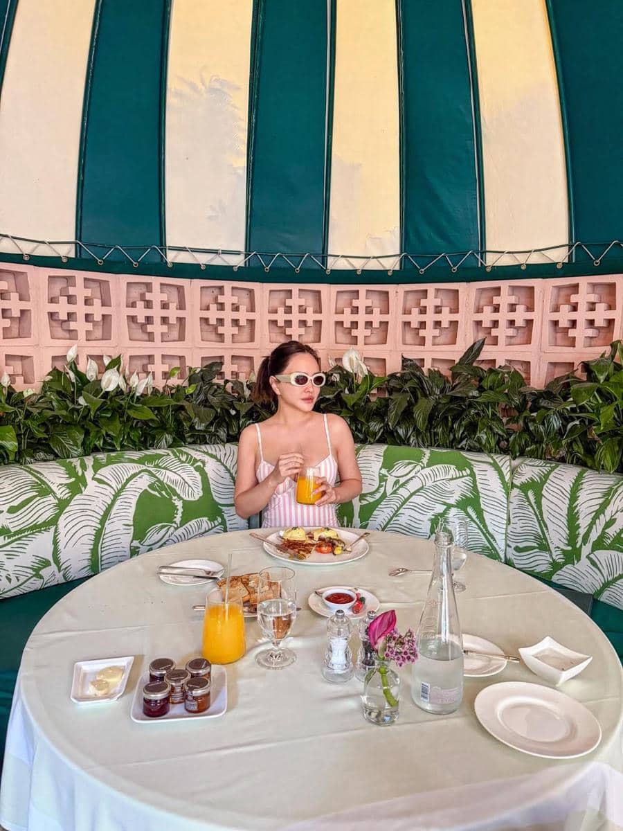 Seorang wanita mengenakan gaun tanpa lengan dan kacamata putih duduk di meja restoran luar ruangan dengan minuman jeruk dan sarapan di depannya.