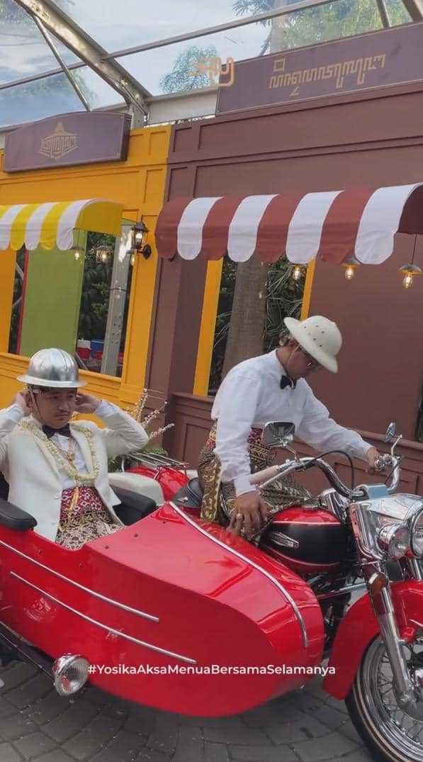 Dua pria mengenakan pakaian adat dan jas putih dengan blangkon modern berpose di atas motor merah dengan sidecar di acara pernikahan.