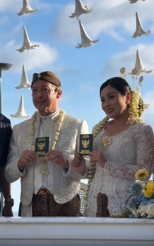 Sepasang pengantin mengenakan busana adat Jawa berwarna putih memegang buku nikah di acara pernikahan dengan latar langit biru.