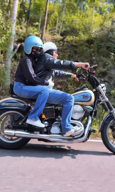 Dua orang mengenakan helm biru dan putih sedang berkendara dengan motor Harley Davidson di jalan dengan latar pepohonan hijau.
