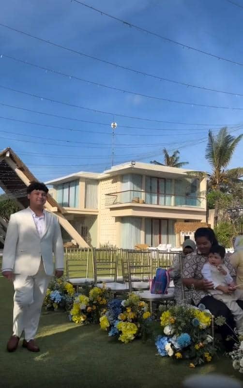 Seorang pria mengenakan setelan jas putih berdiri di taman dengan dekorasi bunga kuning dan biru di depan bangunan modern dua lantai.