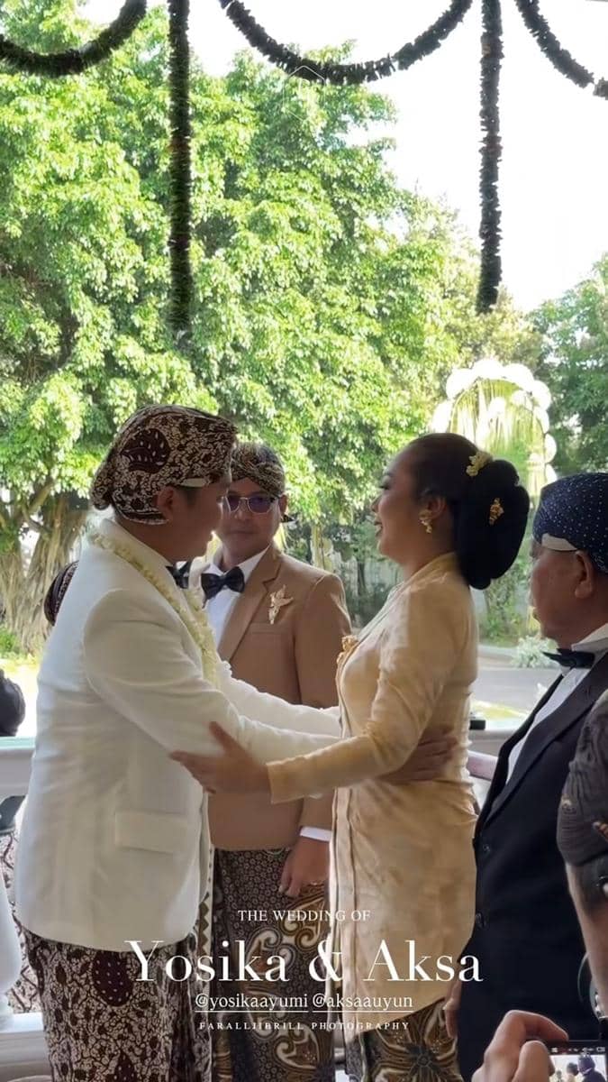 Seorang wanita mengenakan kebaya krem bersalaman dengan pria bersetelan jas putih dan blangkon dalam acara pernikahan adat Jawa.