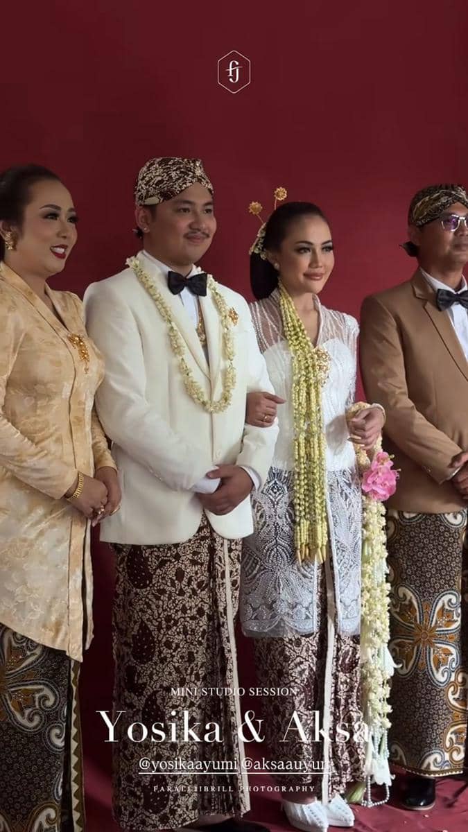 Pasangan pengantin mengenakan busana adat Jawa berwarna putih dan cokelat berpose bersama keluarga di depan latar merah.