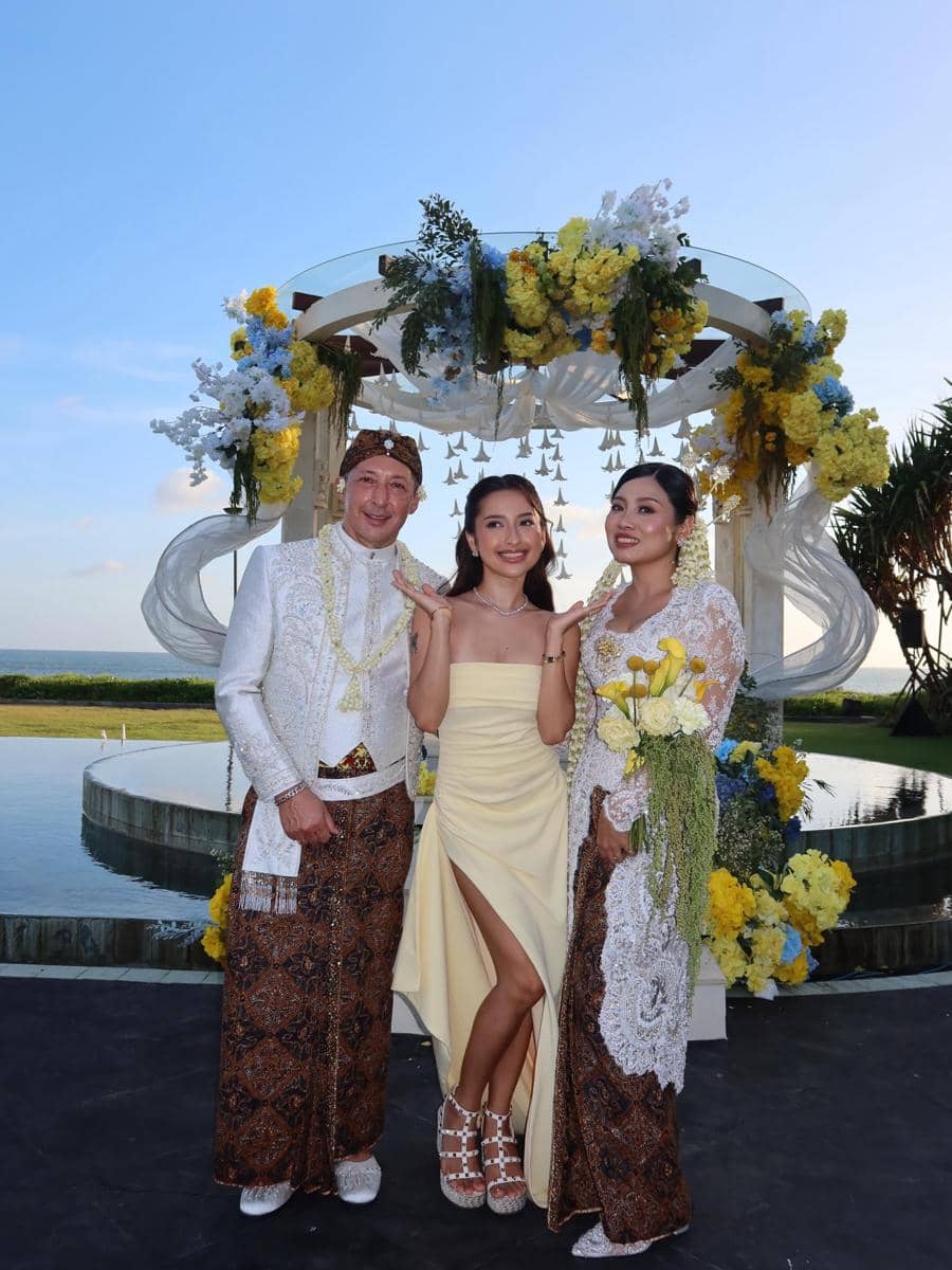 Tiga orang berpose di acara pernikahan dengan latar dekorasi bunga berwarna kuning dan putih di tepi kolam dan langit biru.