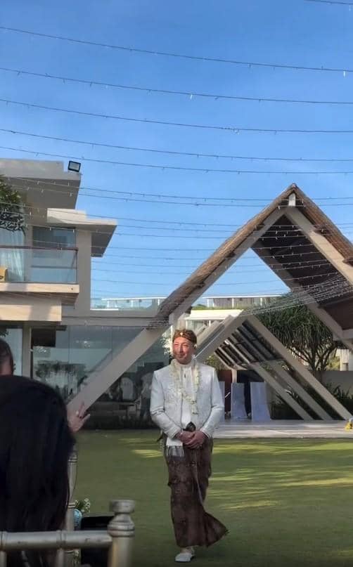 Seorang pria mengenakan busana adat Jawa berwarna putih dan batik cokelat berdiri di area pernikahan outdoor dengan latar bangunan modern.