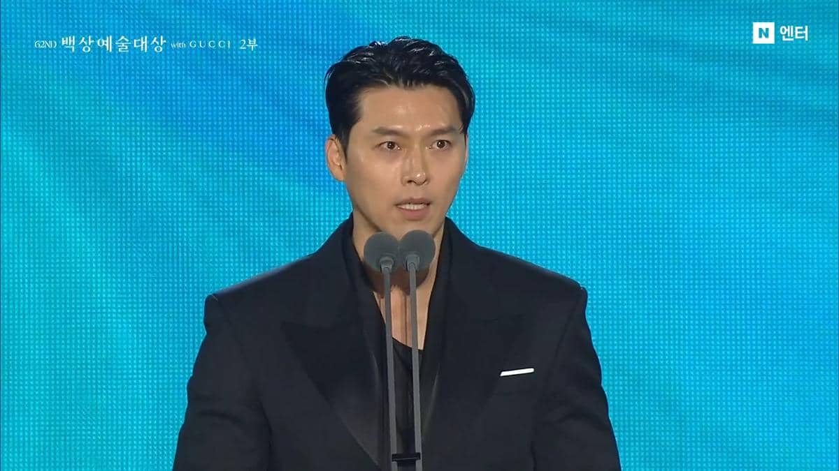 Seorang pria mengenakan setelan jas hitam berbicara di depan mikrofon ganda di panggung acara Baeksang Art Awards 2026.