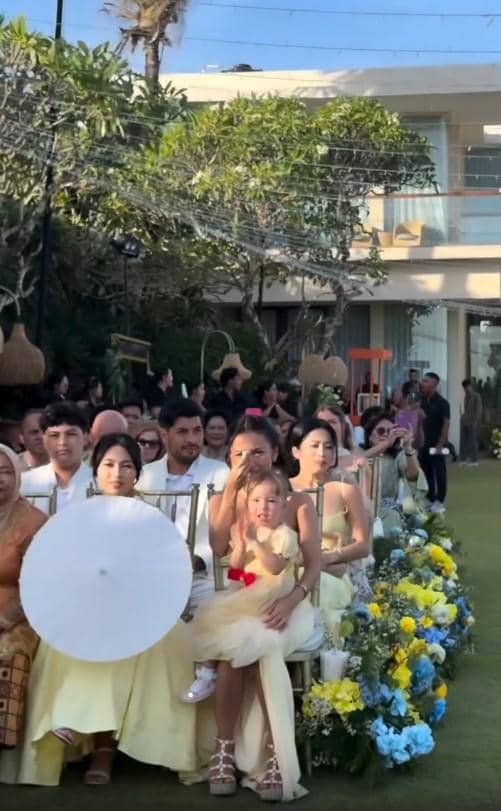 Tamu undangan duduk berderet di acara pernikahan outdoor dengan dekorasi bunga kuning dan biru di siang hari yang cerah.