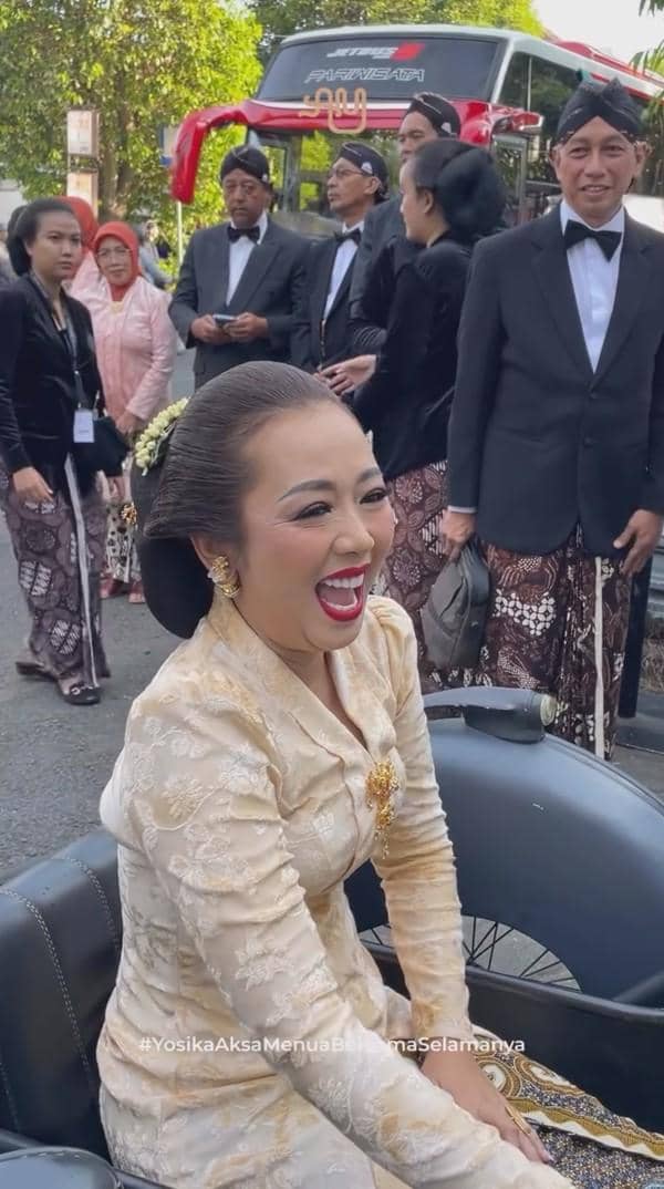 Seorang pengantin wanita mengenakan kebaya krem tersenyum bahagia saat acara akad pernikahan dengan latar tamu berpakaian adat Jawa.
