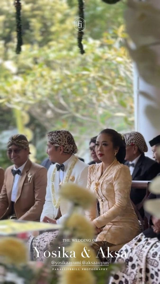 Seorang wanita mengenakan kebaya krem duduk di samping pria berseragam adat Jawa dalam prosesi akad pernikahan di ruang terbuka.