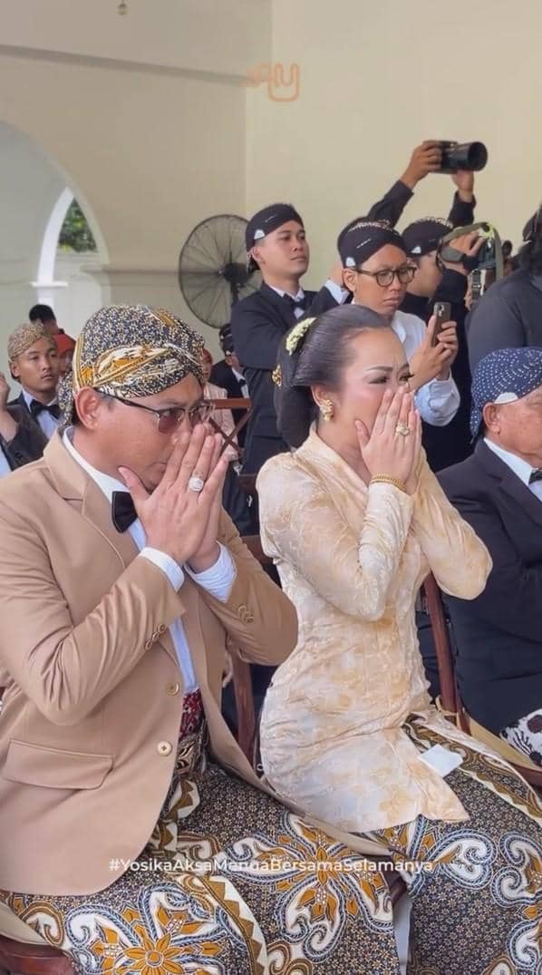 Pasangan pengantin mengenakan busana adat Jawa berwarna cokelat dan krem sedang berdoa khidmat saat prosesi akad pernikahan.