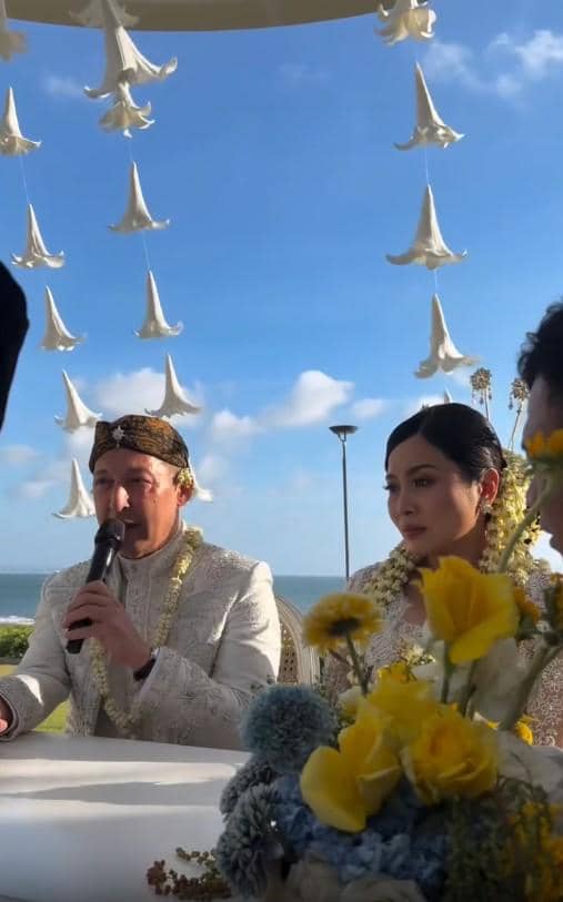 Pasangan pengantin mengenakan busana adat Jawa berwarna putih duduk di meja akad dengan latar laut dan hiasan bunga gantung.