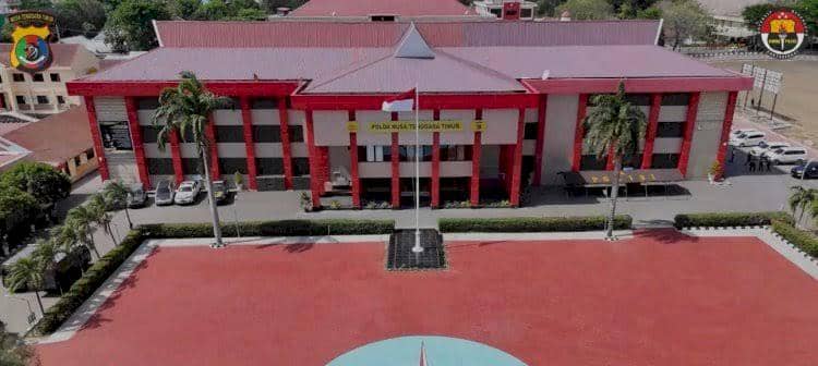 Tampak depan gedung Markas Polda Nusa Tenggara Timur dengan halaman luas berwarna merah dan tiang bendera di tengah area.