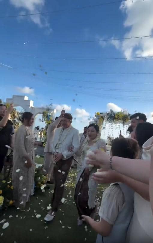 Pasangan pengantin berjalan di tengah tamu yang menaburkan bunga saat upacara pernikahan outdoor dengan langit biru cerah.
